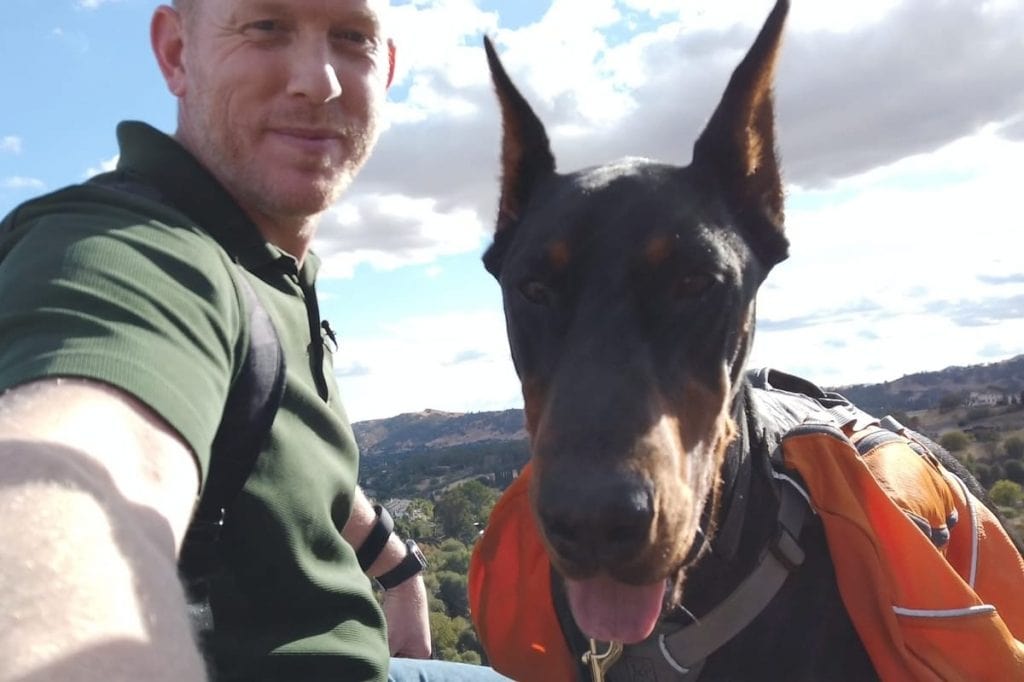 A man and his Doberman at the top of a mountain taking a selfie together.